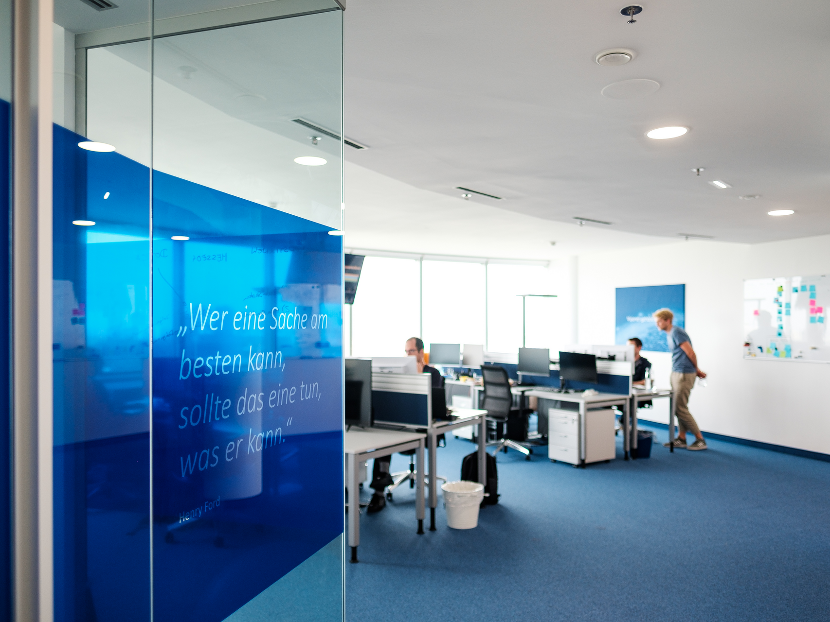 Büroflur mit Glaswand; dahinter mehrere Arbeitsplätze mit Monitoren und Personen im Großraumbüro.