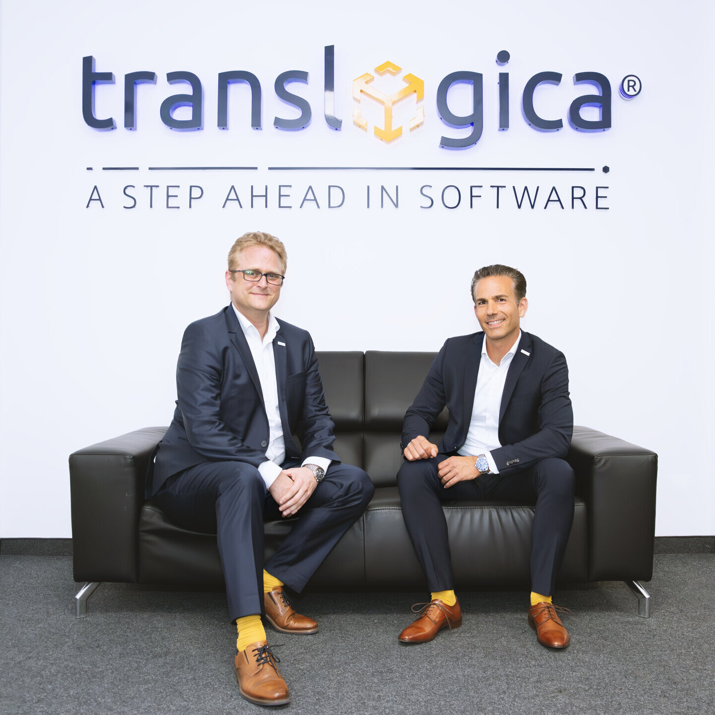 Hansjörg Haller (CEO) und Marc Pfaffenberger (Geschäftsführer) von Translogica sitzen vor dem Firmenlogo auf einem Sofa und sprechen über Softwarelösungen für die Logistik