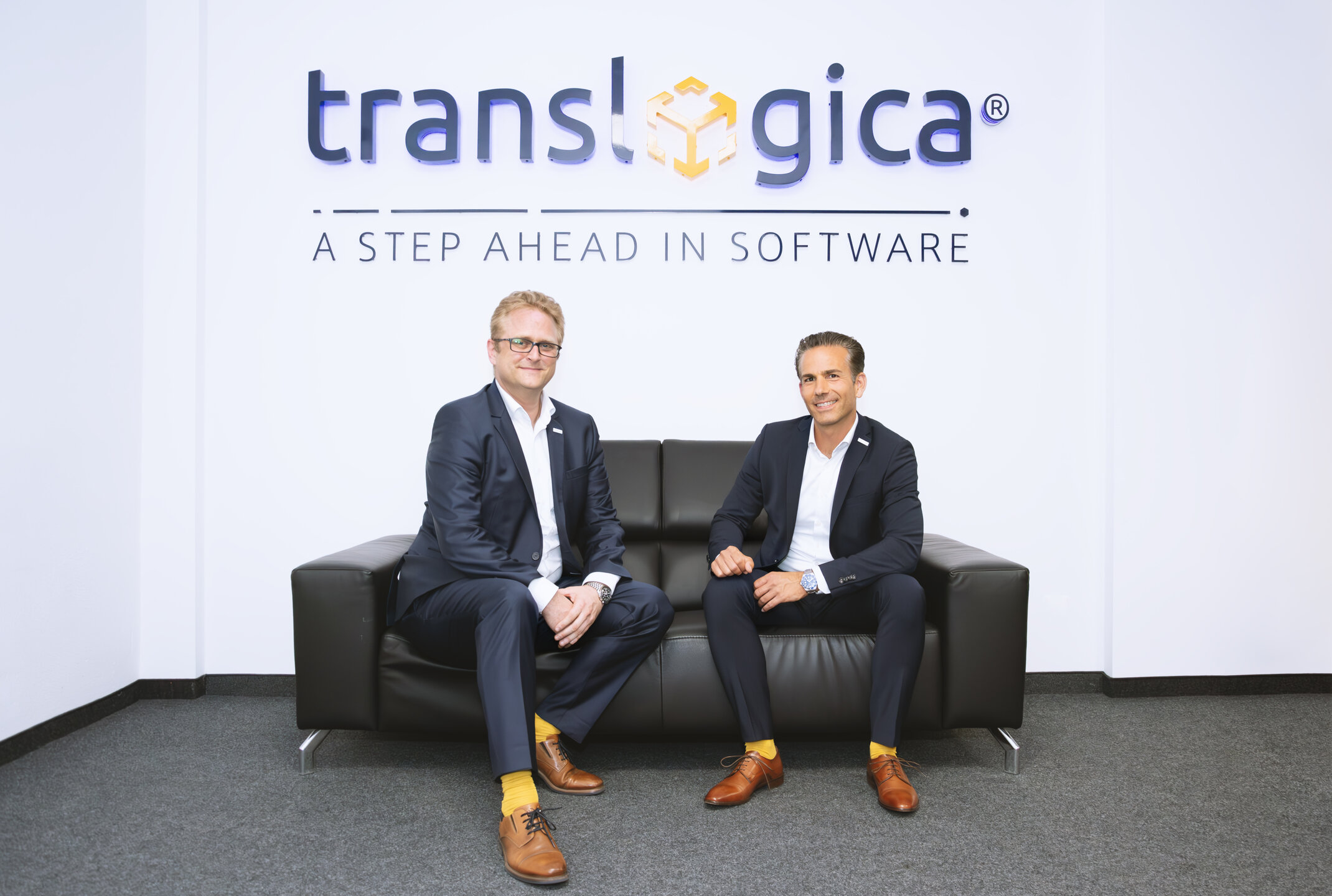 Hansjörg Haller (CEO) und Marc Pfaffenberger (Geschäftsführer) von Translogica sitzen vor dem Firmenlogo auf einem Sofa und sprechen über Softwarelösungen für die Logistik